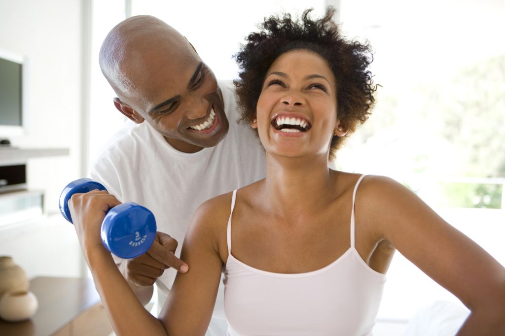 Man smiling at woman lifting weights in living room, woman laughing
