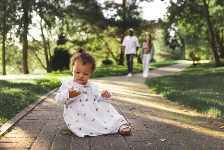 Curious little daughter sitting on park path and exploring nature while parents walk behind. Family lifestyle outdoors, childhood independence, care and safety. Small child at summer day in green park