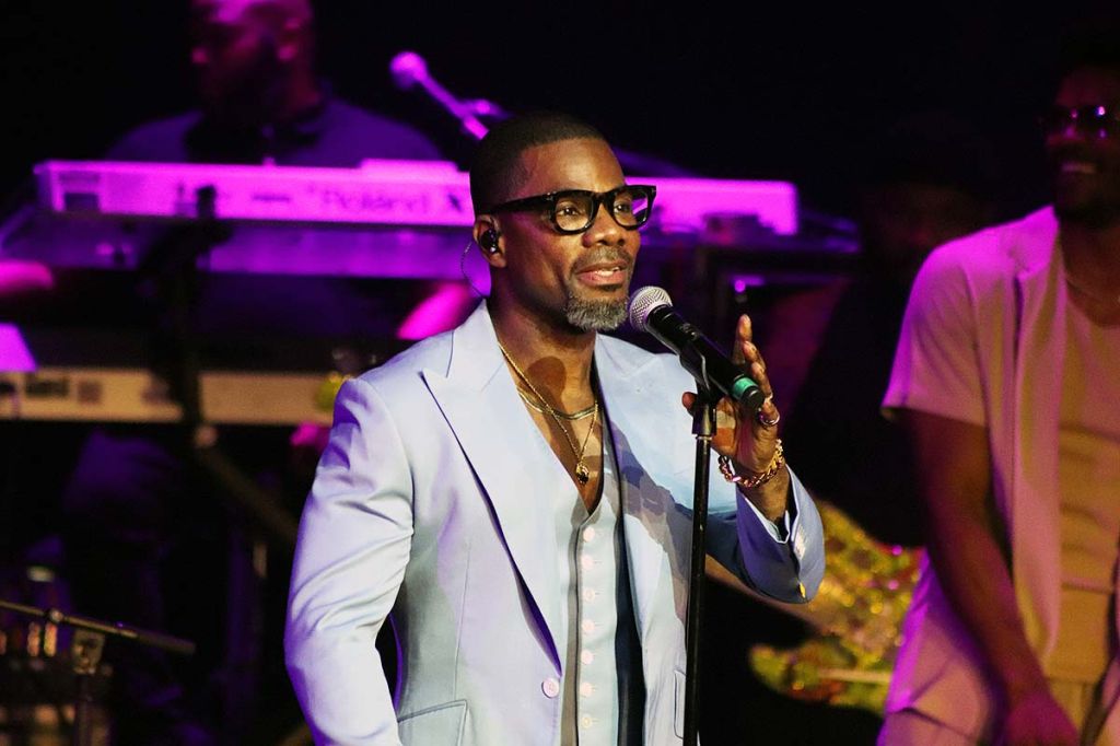 A man wearing glasses and a suit speaking into a microphone on a stage with colorful lighting.