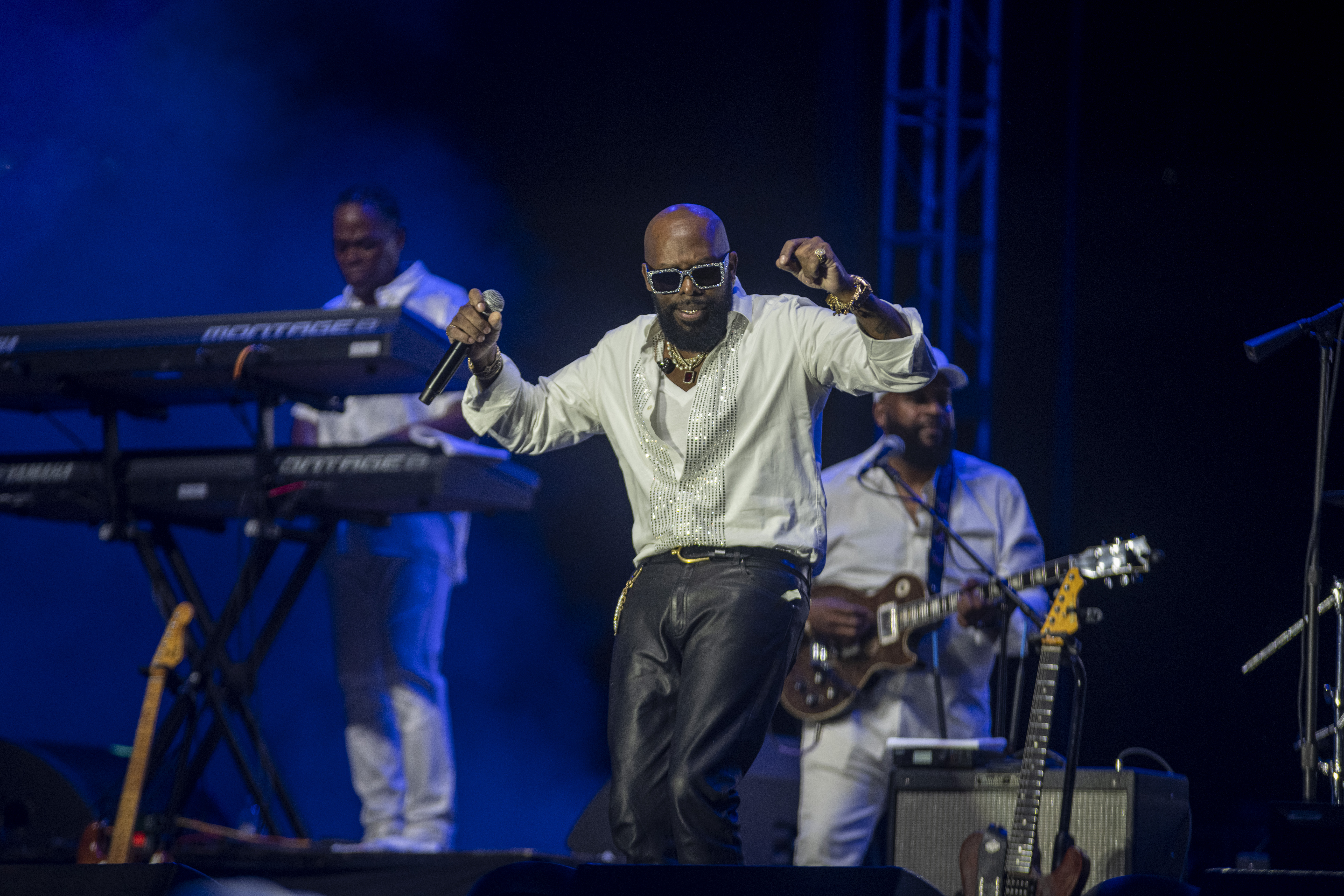 A performer in a white shirt and sunglasses singing and dancing on stage with a band playing behind him.