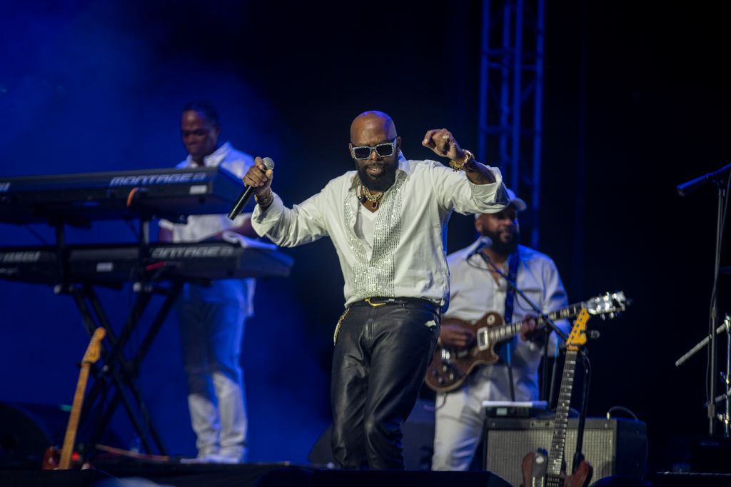 A performer in a white shirt and sunglasses singing and dancing on stage with a band playing behind him.