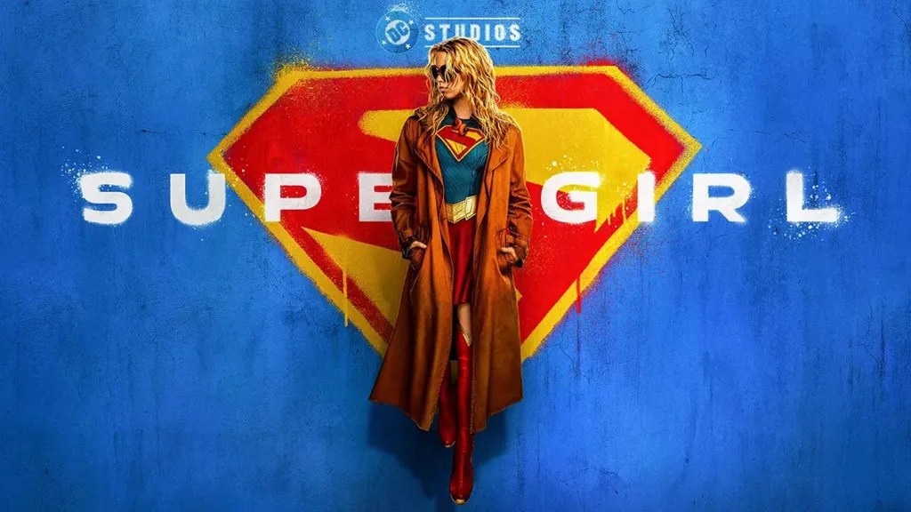 A woman in a red and blue superhero costume stands in front of a blue wall with the "DC Studios" logo, against a backdrop of a large red and yellow Superman-style symbol.