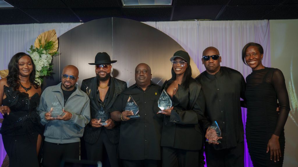 A group of six people, including men and women, posing together and holding awards or trophies. They are dressed formally in suits and dresses, and appear to be at an awards ceremony or event.