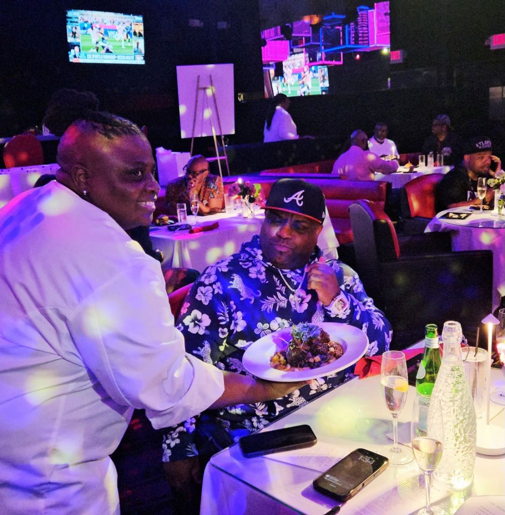 Two people dining at a nightclub with neon lights, one wearing a floral shirt and the other a white suit, enjoying a meal and drinks.