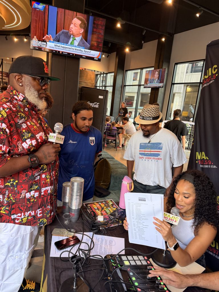 A group of people in a lively setting, with a TV screen displaying a news interview. The people include a man with a colorful shirt, a person wearing a Cape Verde Islands t-shirt, and a woman holding a document.