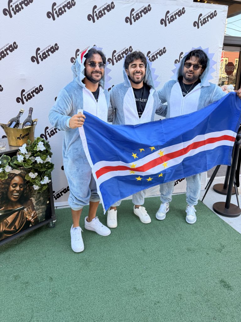 Three men wearing hoodies and sunglasses holding a blue and red flag with stars, standing in front of a Viarae backdrop.