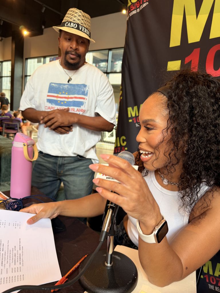 A smiling woman holding a microphone and speaking to a man wearing a Cabo Verde hat and a Cape Verde Islands t-shirt in what appears to be a radio or television studio.