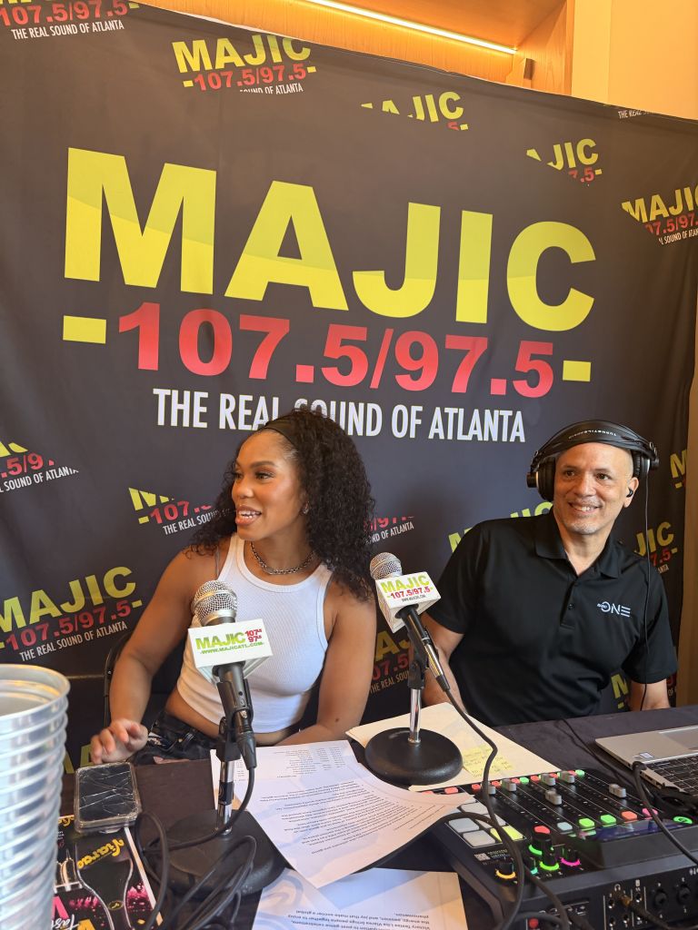 Two people, a woman and a man, holding microphones and standing in front of a large banner for MAJIC 107.5/97.5 radio station in Atlanta.