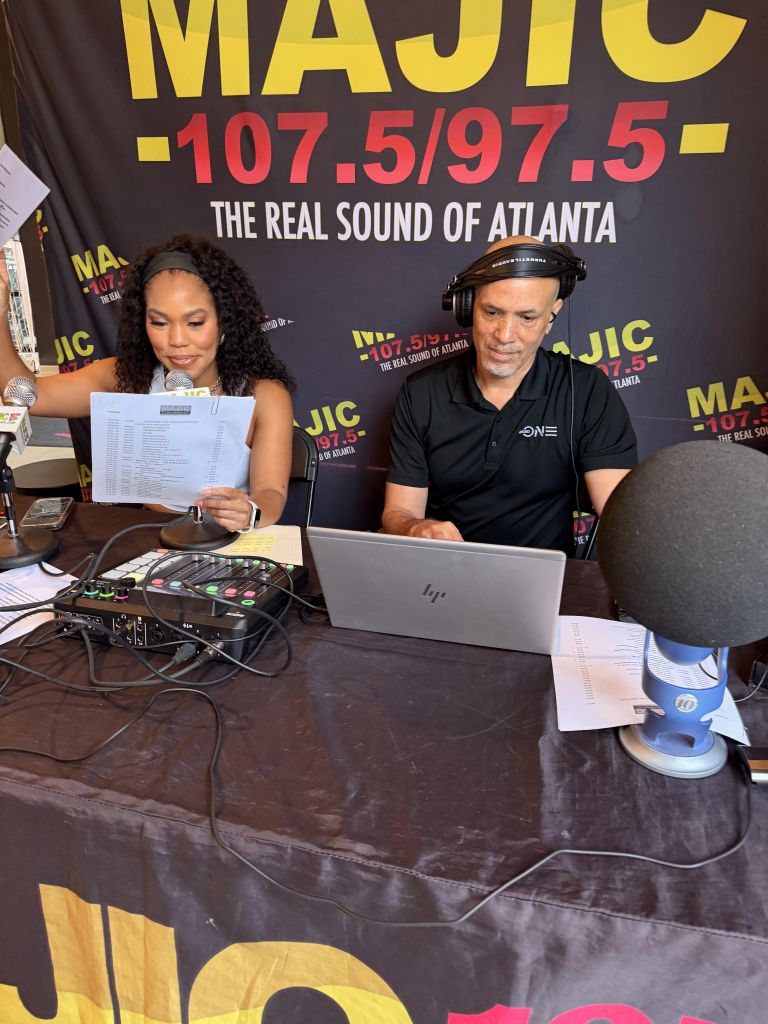 Two people working at a radio station desk, with a laptop, audio equipment, and a large banner for "MAJIC 107.5/97.5 - The Real Sound of Atlanta" visible.