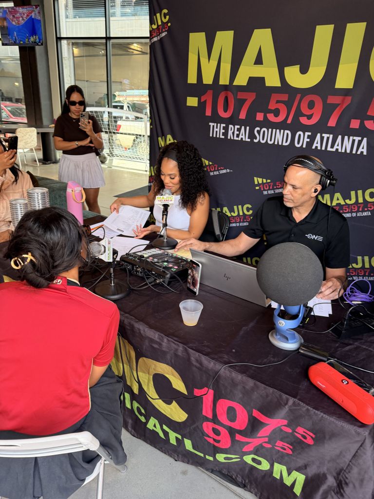 A radio station booth with MAJI 107.5/97.5 branding, featuring several people working at the booth and equipment on the table.