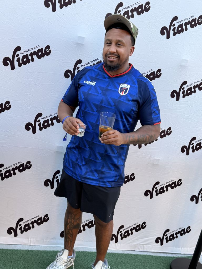 A smiling man wearing a blue soccer jersey with the Capelli logo and holding a glass of beer in front of a Prosecco DOC Viarade banner.