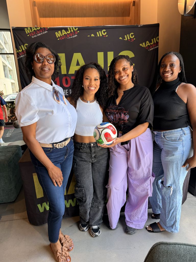 Four young Black women standing in front of a large banner that says "MAJIC 107.5" and displays the station's logo.