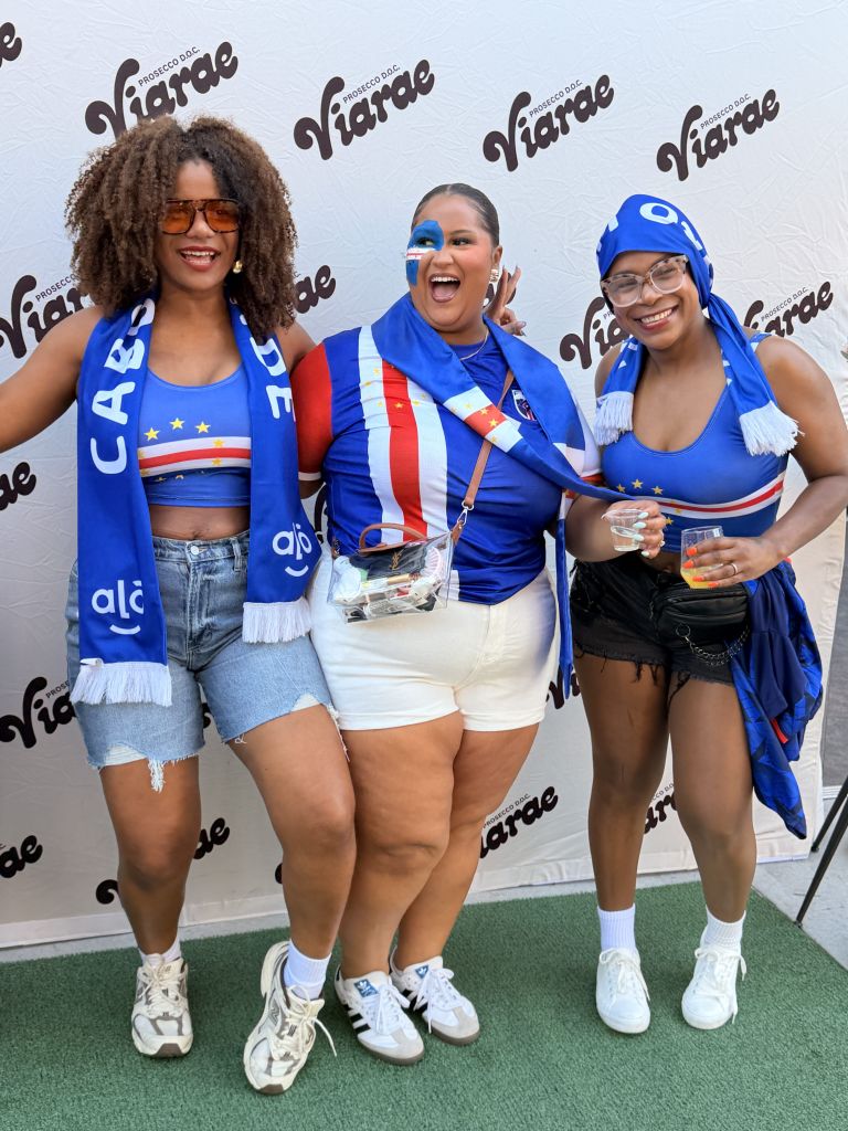 Three smiling women wearing blue and white soccer jerseys, scarves, and hats posing in front of a Viarade backdrop.