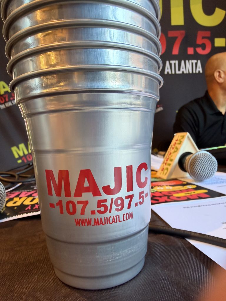A silver metal container with the text "MAJIC 107.5/97.5" and the website "WWW.MAJICATL.COM" printed on it, with a microphone and other radio equipment visible in the background.