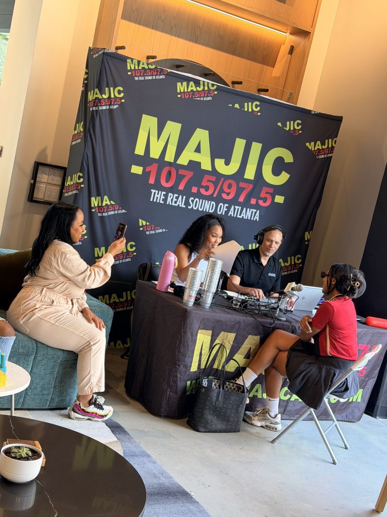 A radio station booth with MAJIC 107.5/97.5 branding, featuring two people operating audio equipment and two others seated nearby in a lounge-like setting.