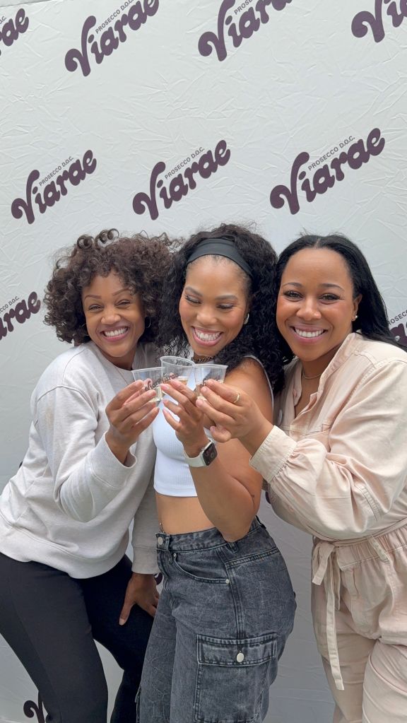 Three smiling women holding glasses of champagne in front of a Prosecco backdrop.