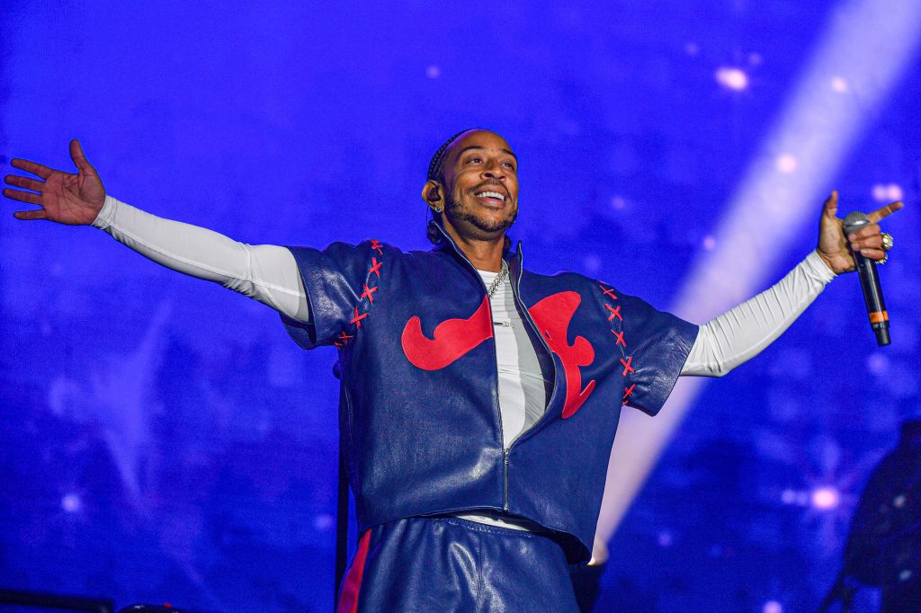 A performer on stage with arms raised, wearing a navy blue and red leather jacket with star designs, smiling and performing before a blue-lit background.