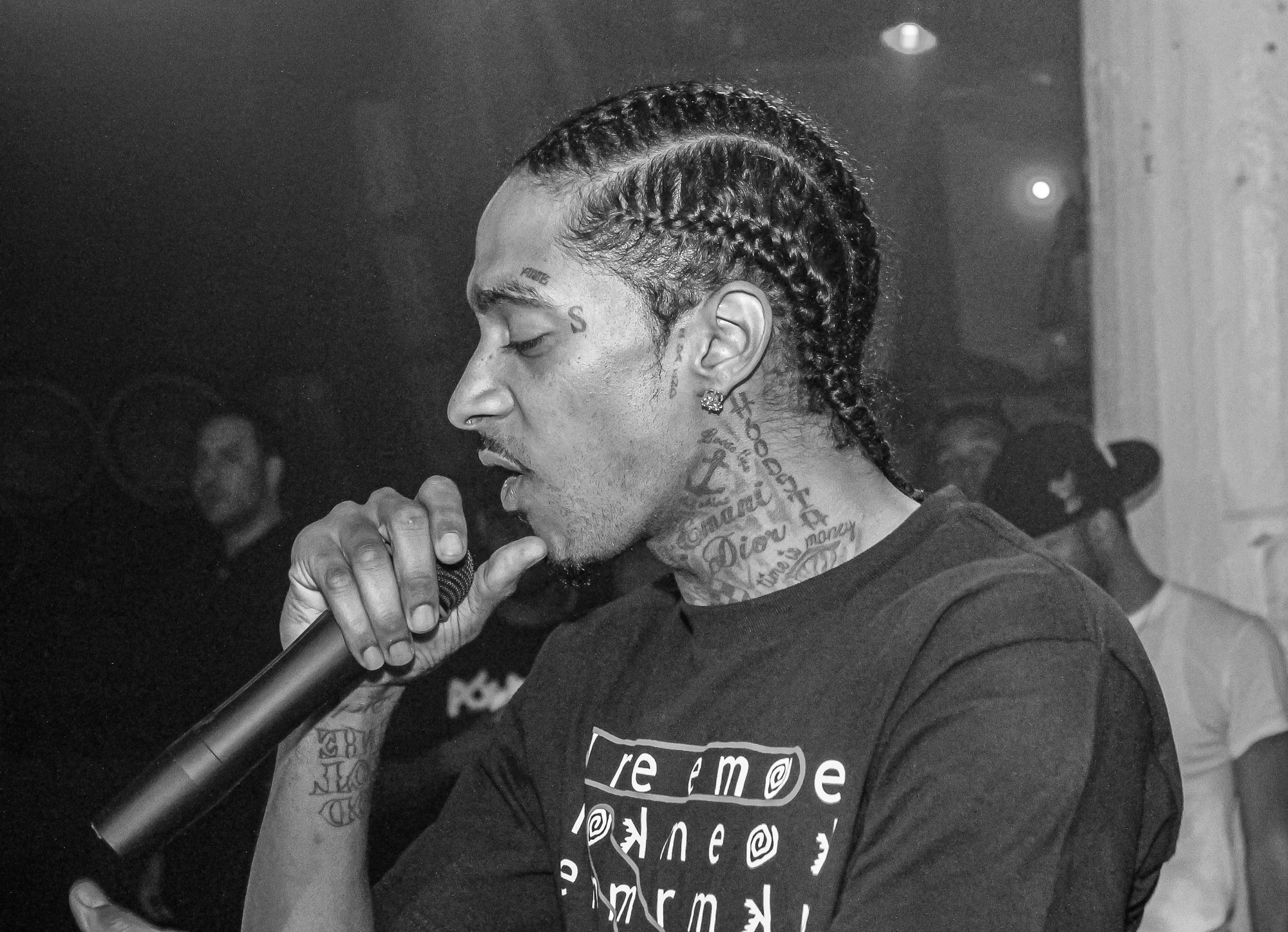 A person with braided hair and tattoos on their face and neck performing on stage, holding a microphone and wearing a shirt with text that reads "Remoe Panes".