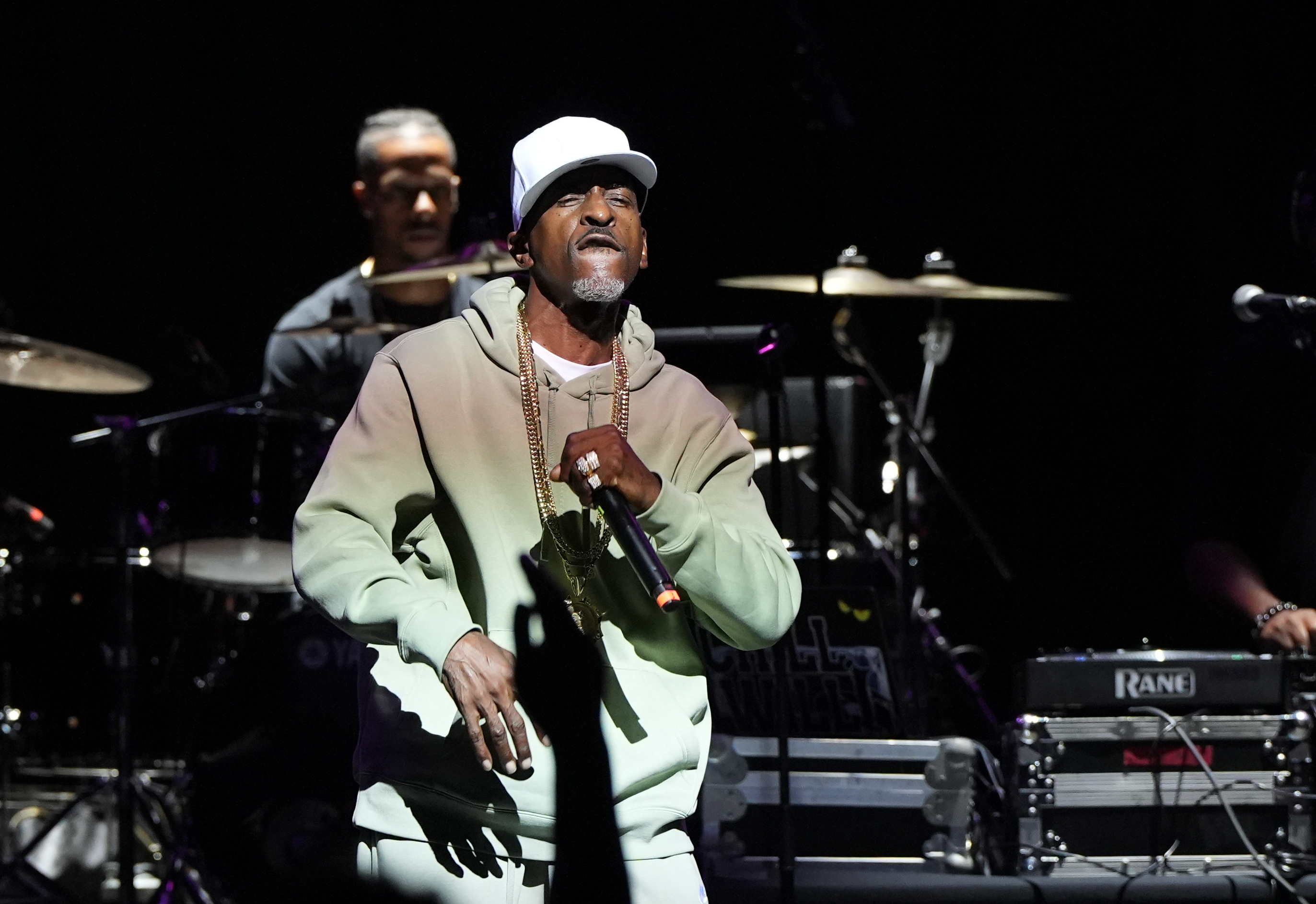A man in a white hat and light-colored clothing performing on stage, with a drummer visible in the background.