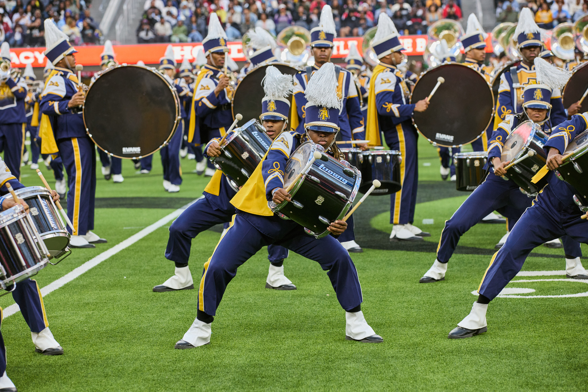 A marching band performs on a football field, with drummers and musicians in navy blue and gold uniforms holding instruments.
