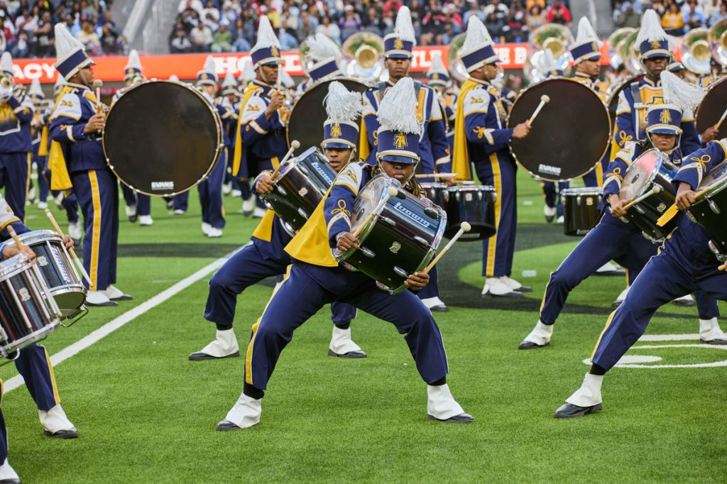 A marching band performs on a football field, with drummers and musicians in navy blue and gold uniforms holding instruments.