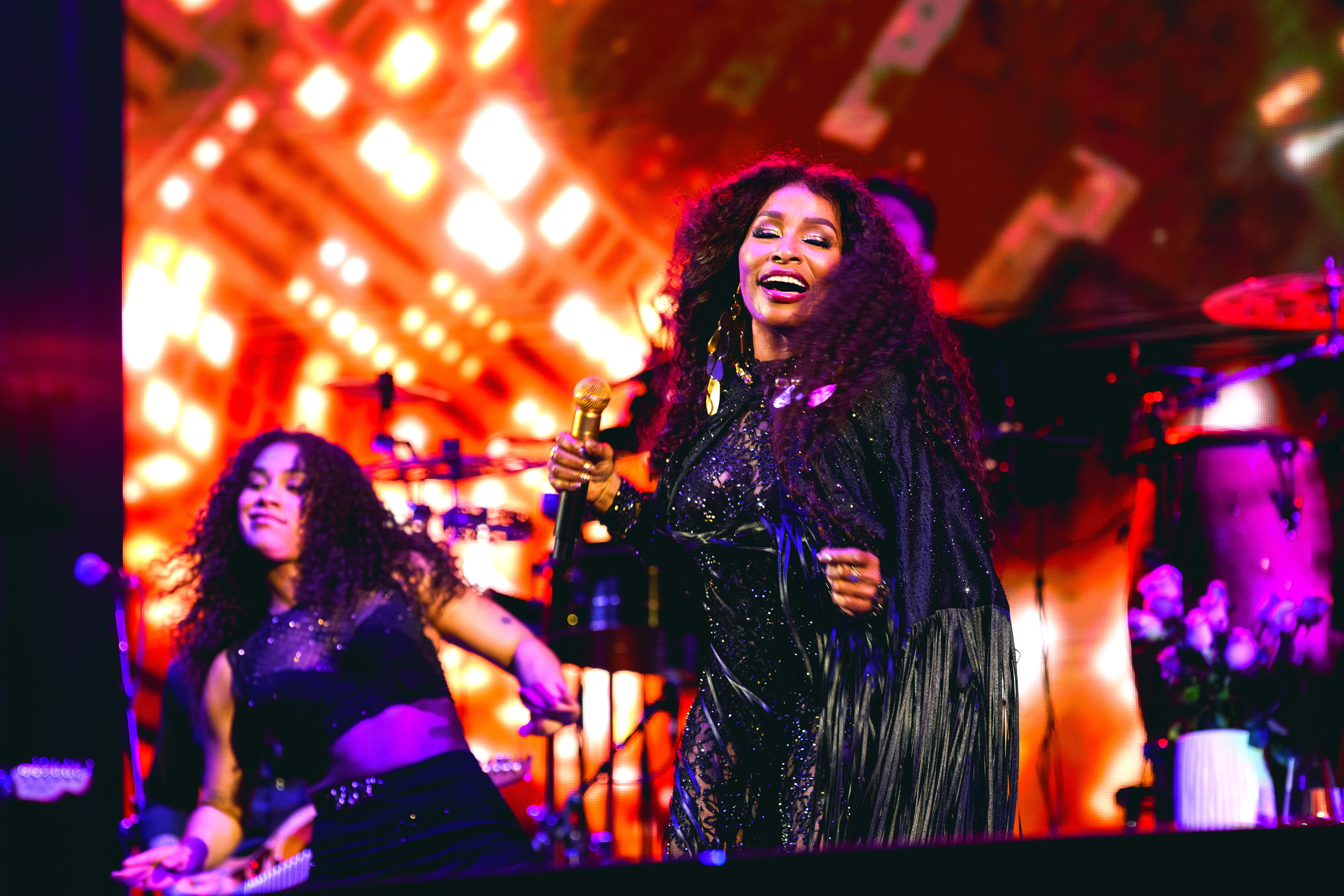 A woman with curly hair performing on stage, wearing a sparkling black dress and cape, singing into a microphone with a bright, joyful expression.