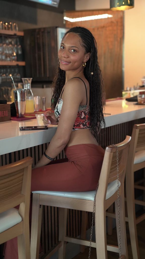 A smiling woman with braided hair sitting at a bar, wearing a patterned top and holding a drink.
