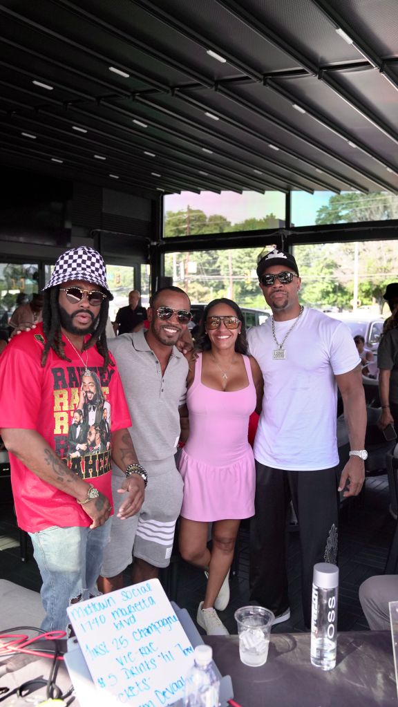A group of four people, including two men and two women, posing together outdoors. One man is wearing a red t-shirt with a graphic design, and the other man is wearing a white t-shirt. The women are wearing a pink dress and a pink tank top. They appear to be at some kind of event or gathering.