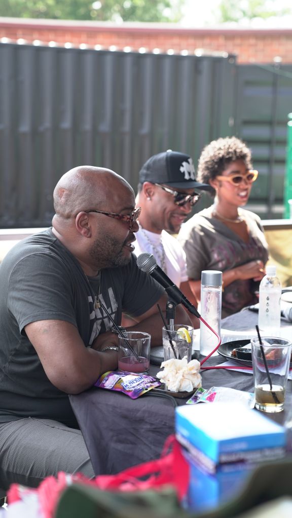 Three people, two men and one woman, sitting at a table with microphones, drinks, and other items. The man in the center is wearing a New York Yankees baseball cap.