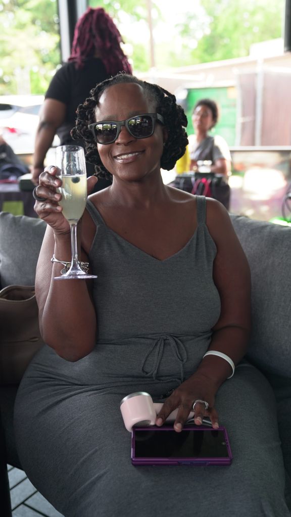 A smiling woman wearing sunglasses and holding a glass of lemonade while sitting on a couch and using a mobile device.
