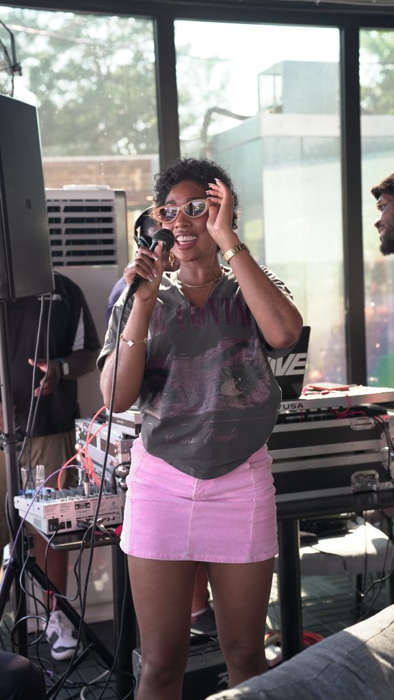 A smiling woman wearing sunglasses and holding a microphone on stage, with audio equipment and a window visible in the background.