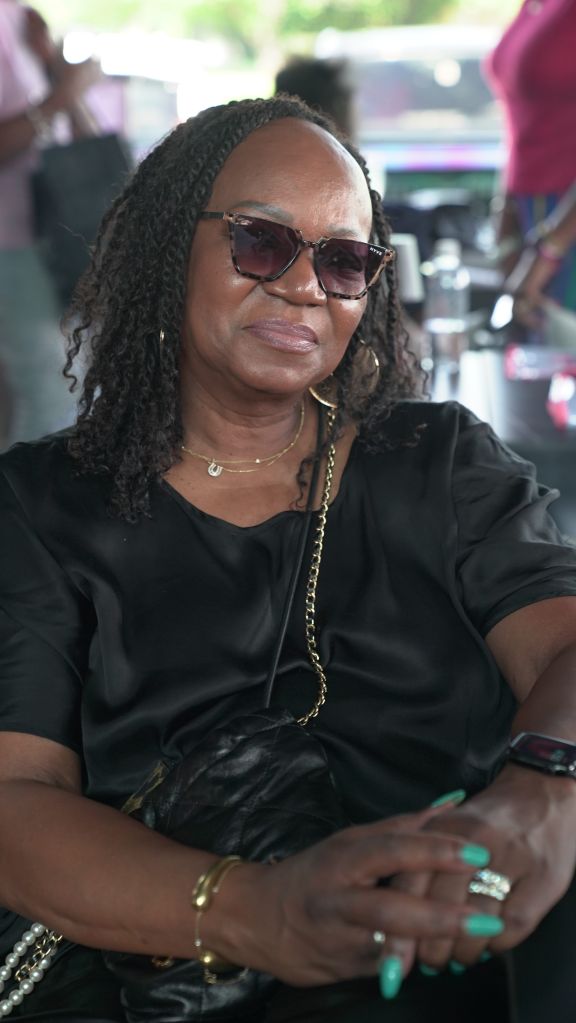 A woman with braided hair wearing sunglasses and a black jacket, sitting in what appears to be a vehicle or public transit.