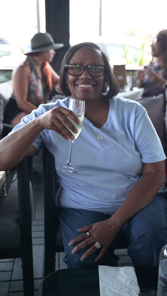 A smiling older woman wearing glasses and a white t-shirt holding a glass of champagne.