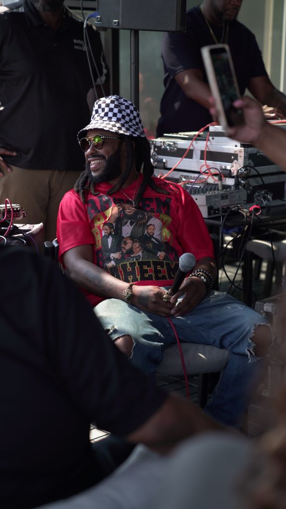 A person wearing a red shirt with a graphic design, a checkered bucket hat, and sunglasses, sitting in front of audio equipment.
