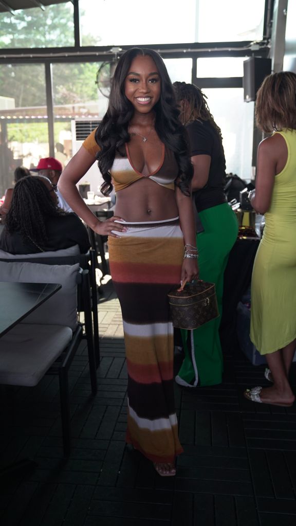 A smiling woman wearing a crop top and striped skirt stands in a room with other people.