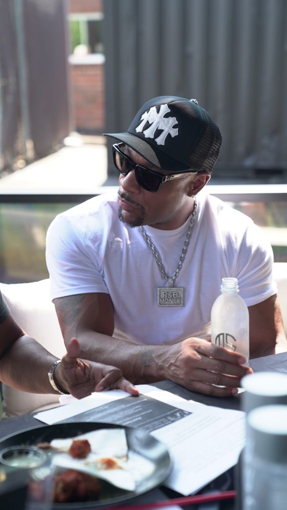 A man wearing a black hat with a white logo, sunglasses, and a white t-shirt with a logo, sitting at a table with a bottle and papers in front of him.