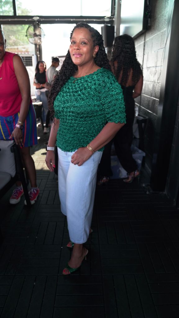 A woman wearing a green crocheted top and white pants stands in a setting with other people in the background.