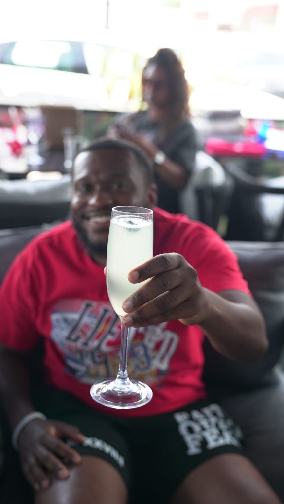A person in a red shirt holding a glass of what appears to be a mixed drink, smiling and enjoying themselves in a casual setting.