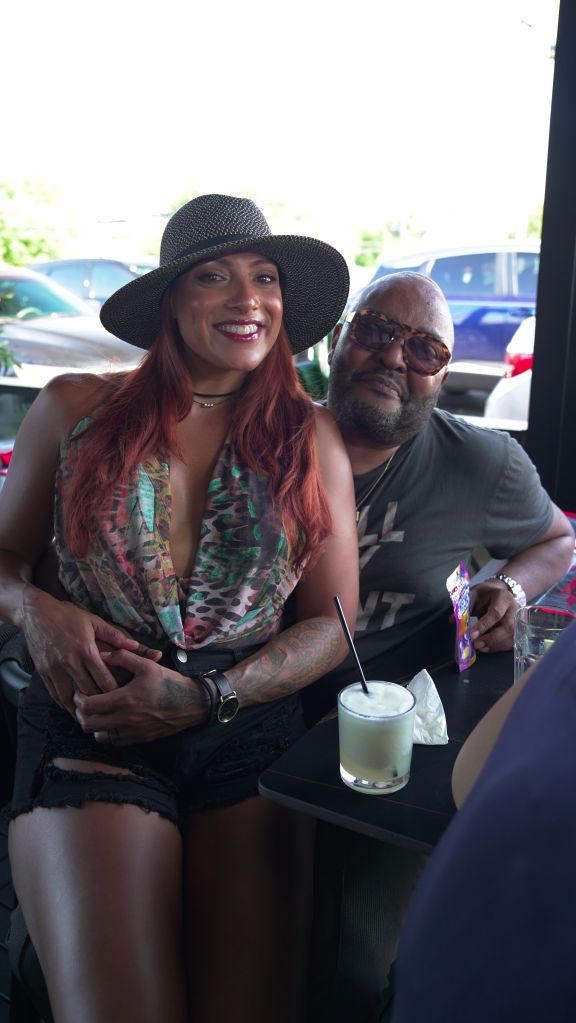 A smiling woman with red hair wearing a patterned top and a large black hat, sitting next to a man with sunglasses and a beard holding a drink.
