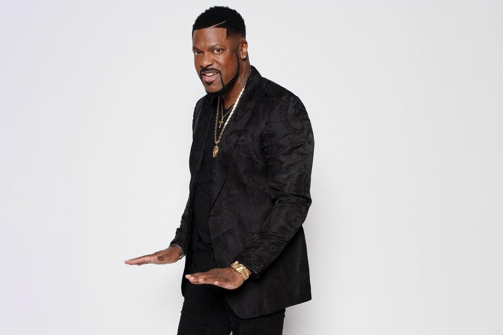 A Black man wearing a black suit and gold jewelry stands with his arms outstretched in a confident pose against a plain white background.