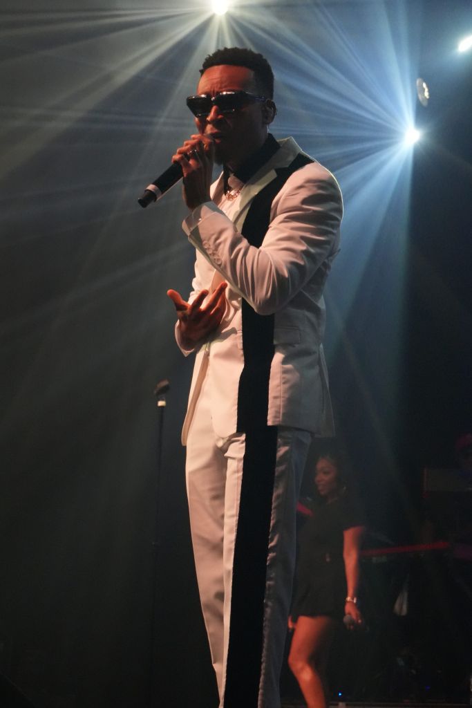 A man in a white suit and sunglasses performing on stage, with bright lights shining behind him.