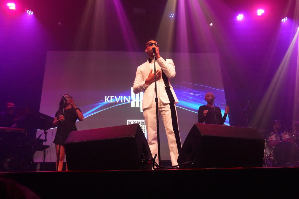 A performer in a white suit singing on stage with a microphone, surrounded by colorful stage lighting and a large "KEVIN" display behind them.