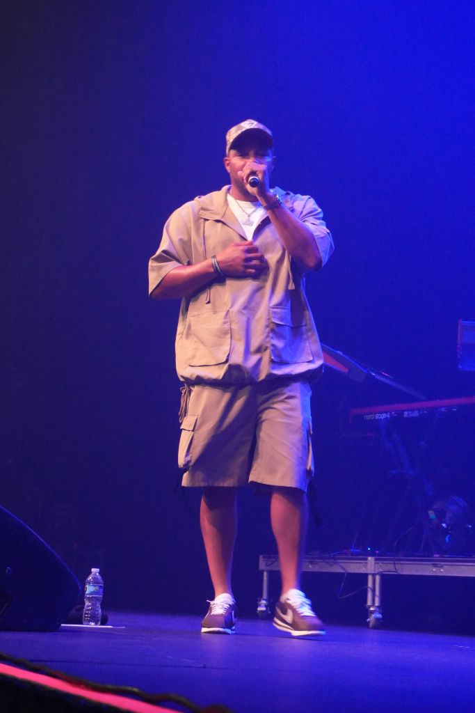A person wearing a beige jacket and shorts performs on a stage under blue lighting, holding a microphone.