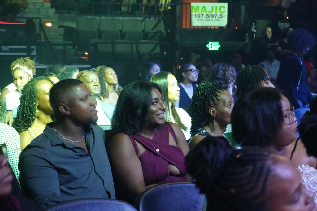 A crowded indoor event with people wearing formal attire. The MAJIC 107.5 logo is visible on a screen in the background.