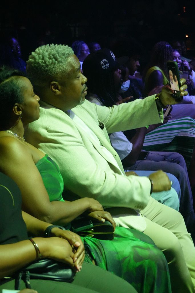 A person in a bright green outfit sitting in a crowd, holding a smartphone and taking a selfie.
