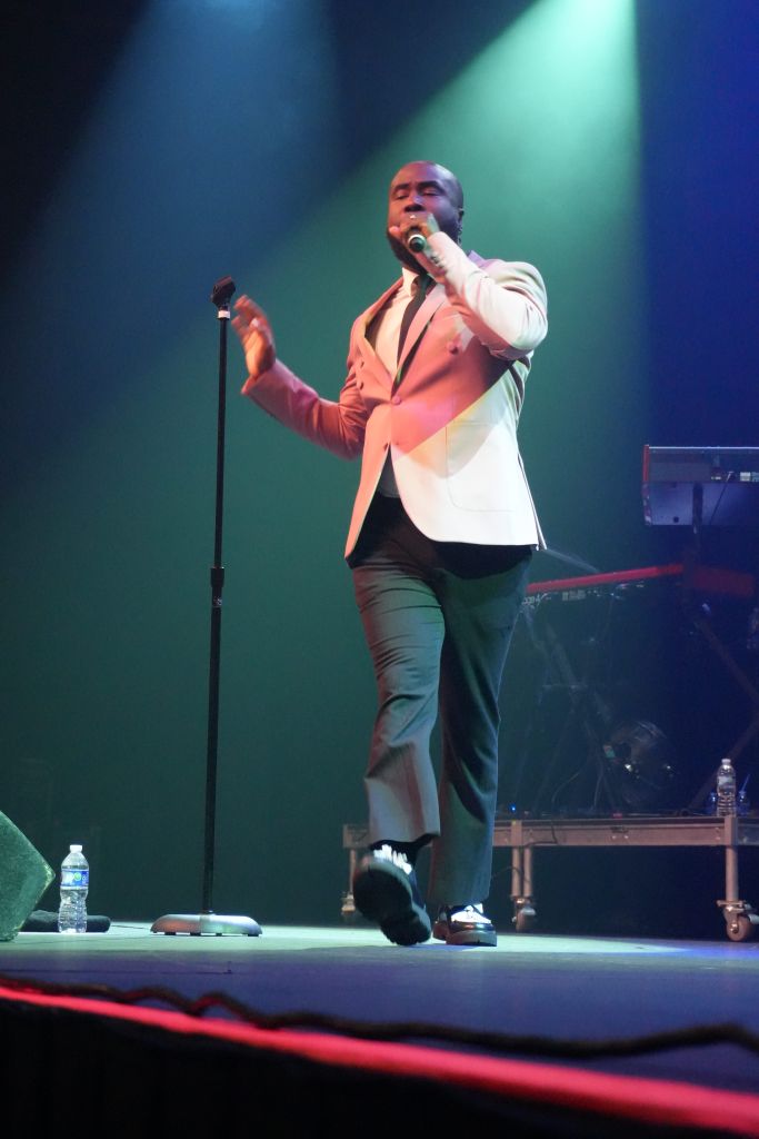 A man in a red suit performing on stage, holding a microphone and gesturing with his hands.