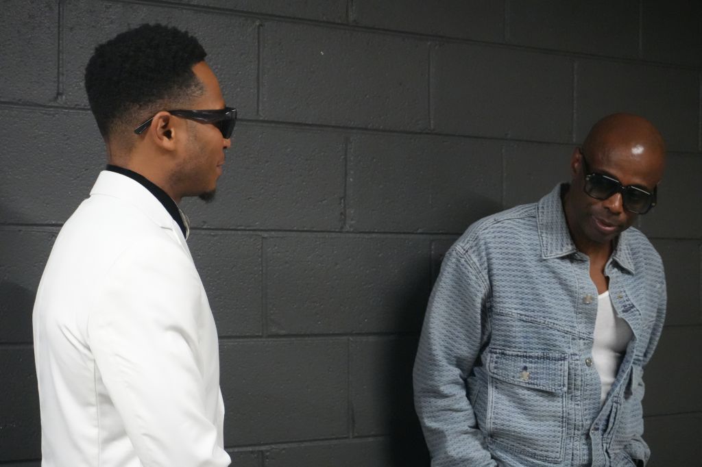 Two men wearing sunglasses standing against a dark wall