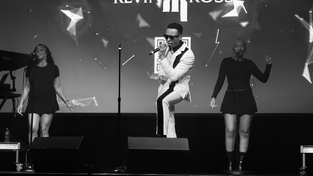 Three performers on stage, one in a white suit singing into a microphone, flanked by two dancers in black outfits.