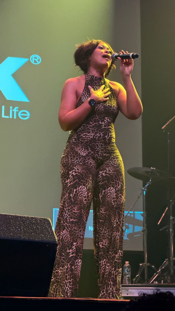 A woman in a leopard print jumpsuit singing into a microphone on stage, with a large "Life" logo visible behind her.