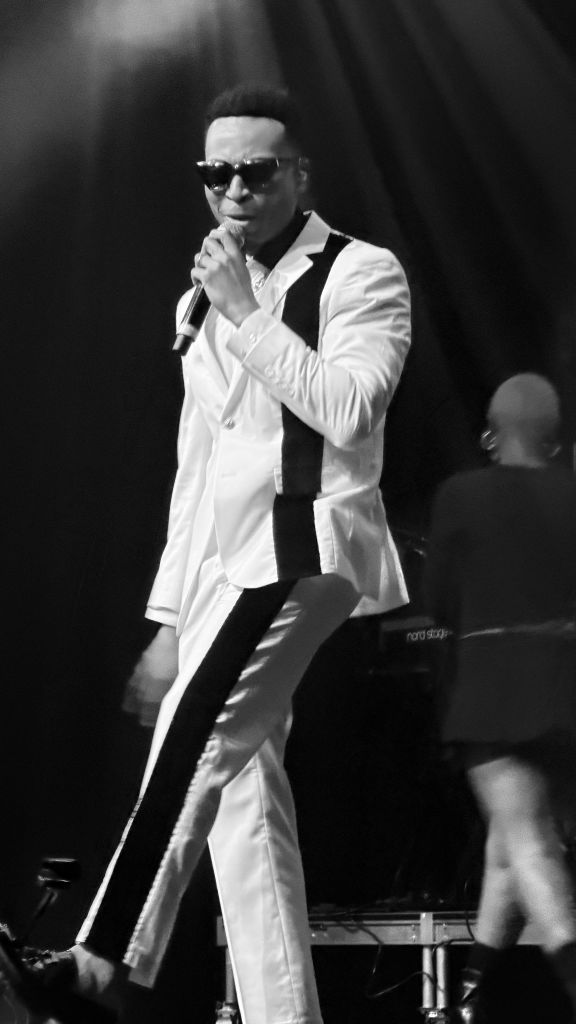 A man in a white suit and sunglasses performing on stage, holding a microphone.